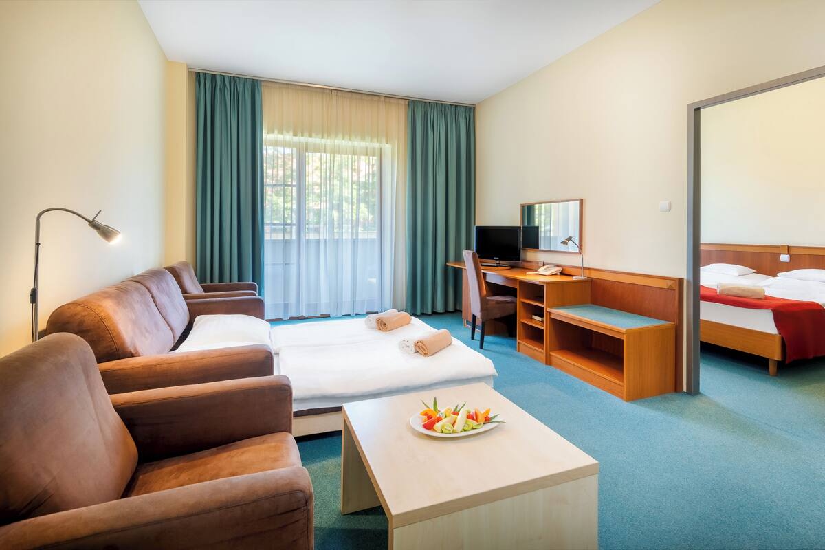 Une chambre d’hôtel comprenant un lit, un bureau, une chaise, un canapé et une table à manger avec des fruits.