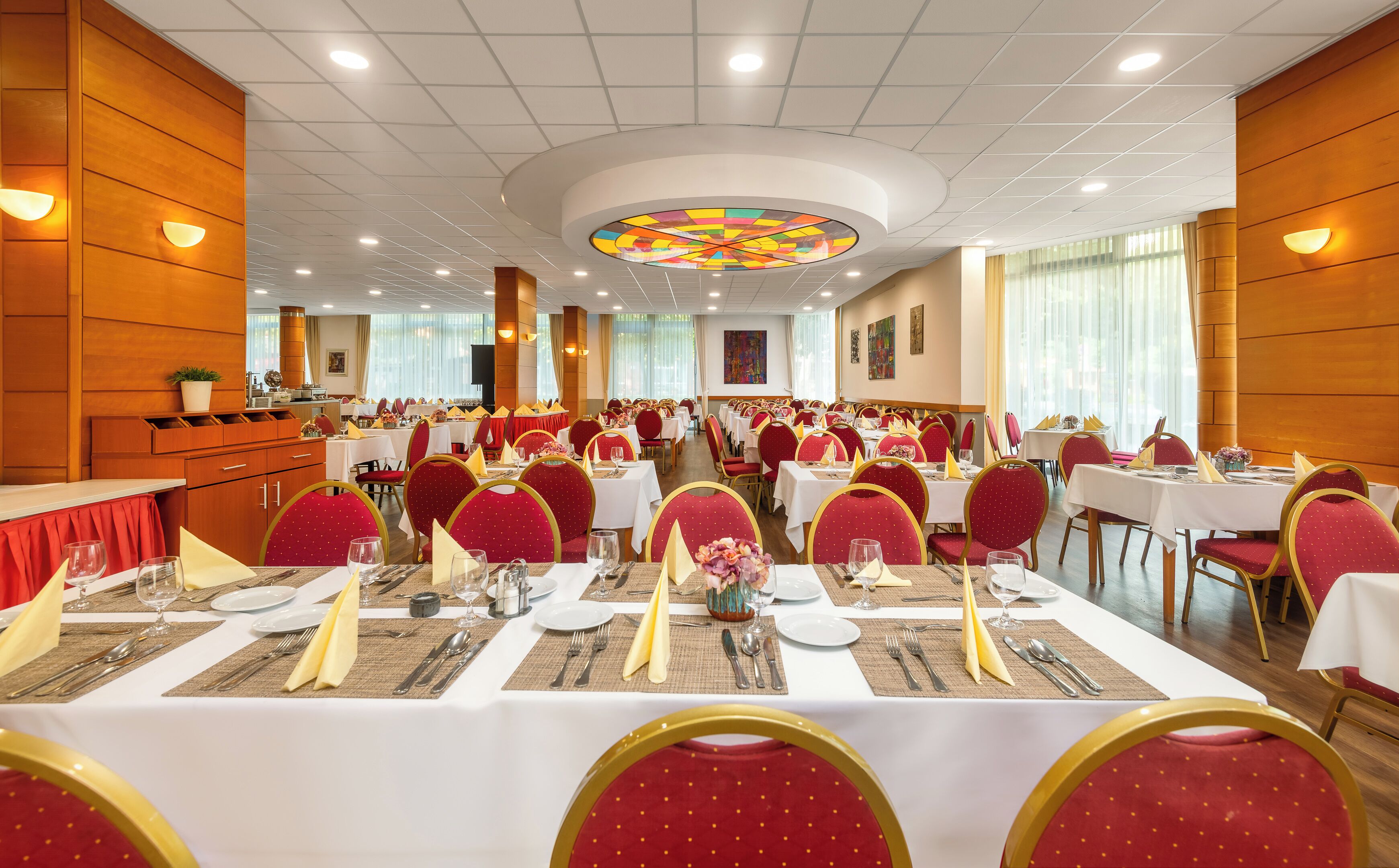 A banquet hall with round tables, red upholstered chairs, and a central stained glass window.