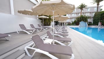 Zona junto a la piscina con tumbonas y sombrillas, una piscina azul cristalina y un edificio con balcones al fondo.