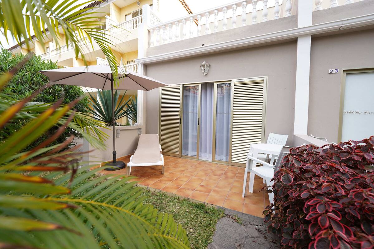 A courtyard with a white lounge chair, a table, and an umbrella. There are plants and a building with a balcony in the background.