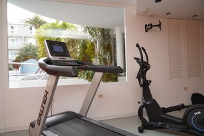 Ein Heimfitnessraum mit Laufband und Ellipsentrainer, ein Fenster mit Blick auf einen Balkon und viel Grün, sowie an der Wand befestigte Fitnessgeräte.