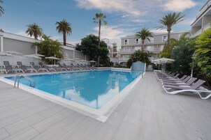 A swimming pool with lounge chairs and umbrellas, surrounded by palm trees and a building with balconies.