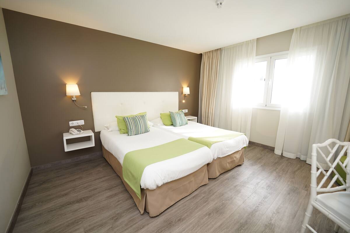 A double bed with green and white bedding, a bedside table with a lamp, and a window with curtains.