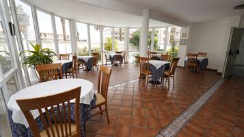 Un amplio comedor interior con varias mesas y sillas, ventanas grandes que ofrecen vistas al exterior y piso de baldosas.