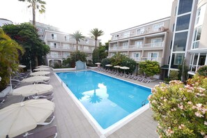 Ein Swimmingpool, umgeben von Liegestühlen und Sonnenschirmen, mit einem Gebäude im Hintergrund, das Balkone und ein Fliesenmuster aufweist.