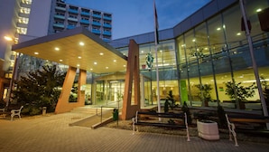 Um edifício moderno com fachada de vidro, entrada destacada com galeria coberta e um mastro de bandeira.