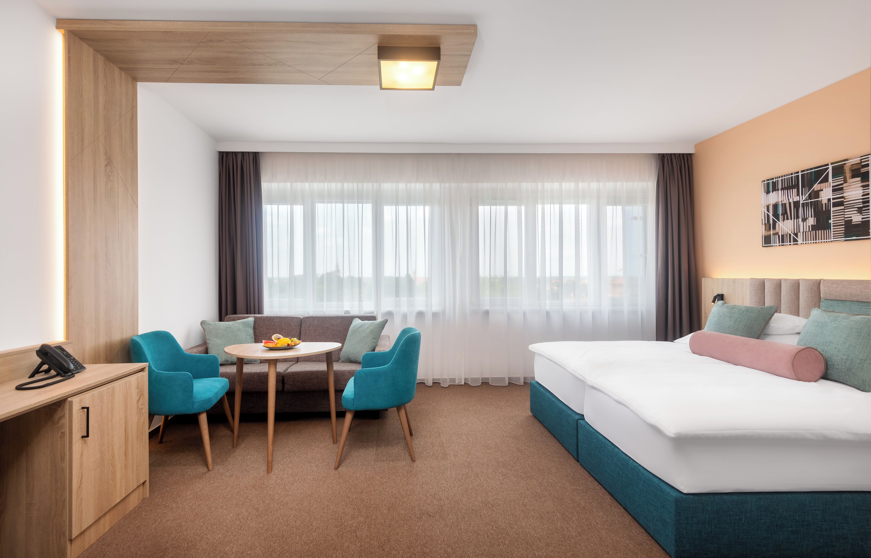 A hotel room with a bed, a desk, a chair, a small table, and a window with curtains.
