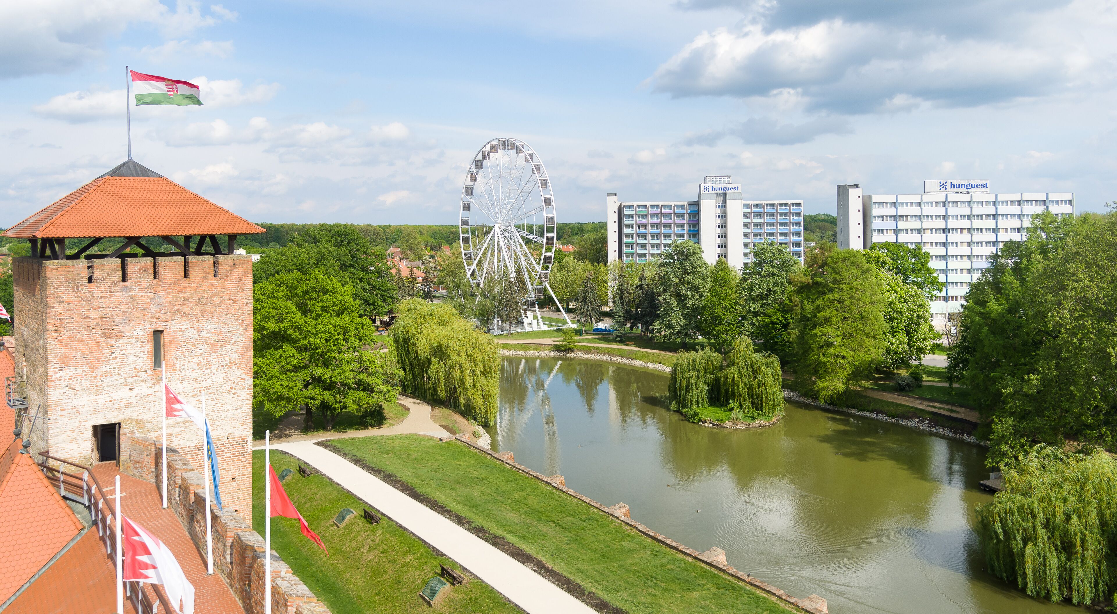 A historic tower with a flag, a river with a path, a Ferris wheel, and modern buildings in the background.