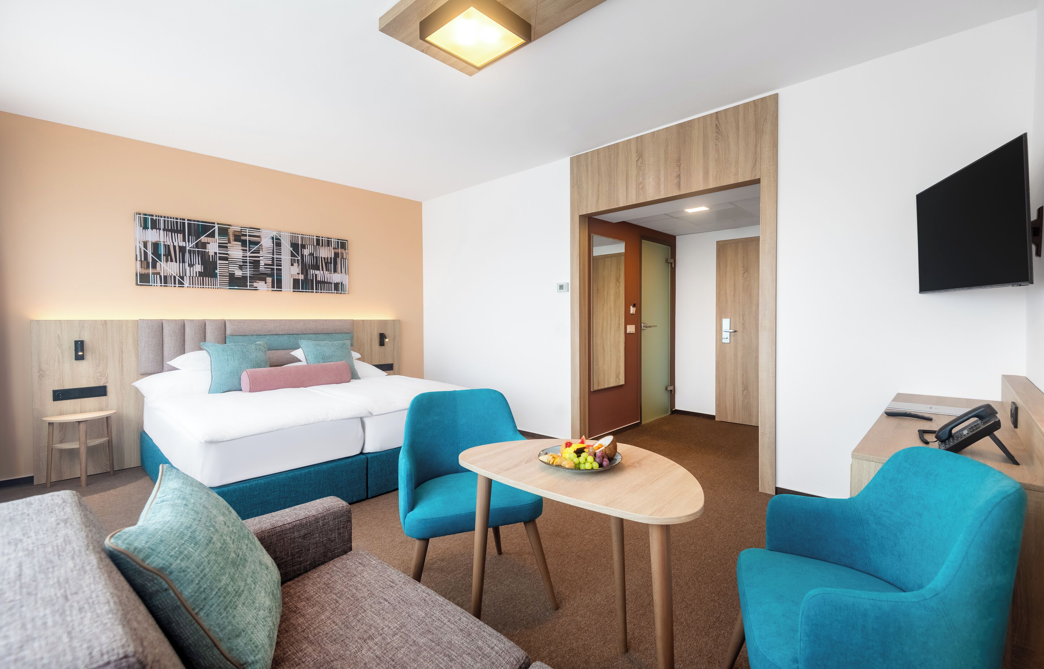 A hotel room with a bed, two blue armchairs, a small table with fruit, a TV, and a wooden door.