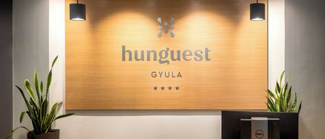 A reception desk with a wooden panel displaying the name "hunguest" and "GYULA" with a star rating of five stars. There are two potted plants on either side of the desk.