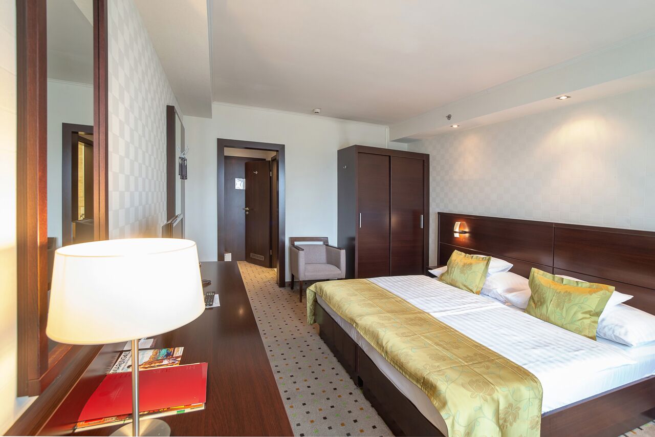 A hotel room with a large bed, a desk with a lamp, a chair, and a wardrobe.