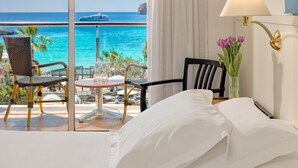 Habitación de hotel con cama, dos sillas, una mesita con botellas, un jarrón con flores y vista al océano y un barco.