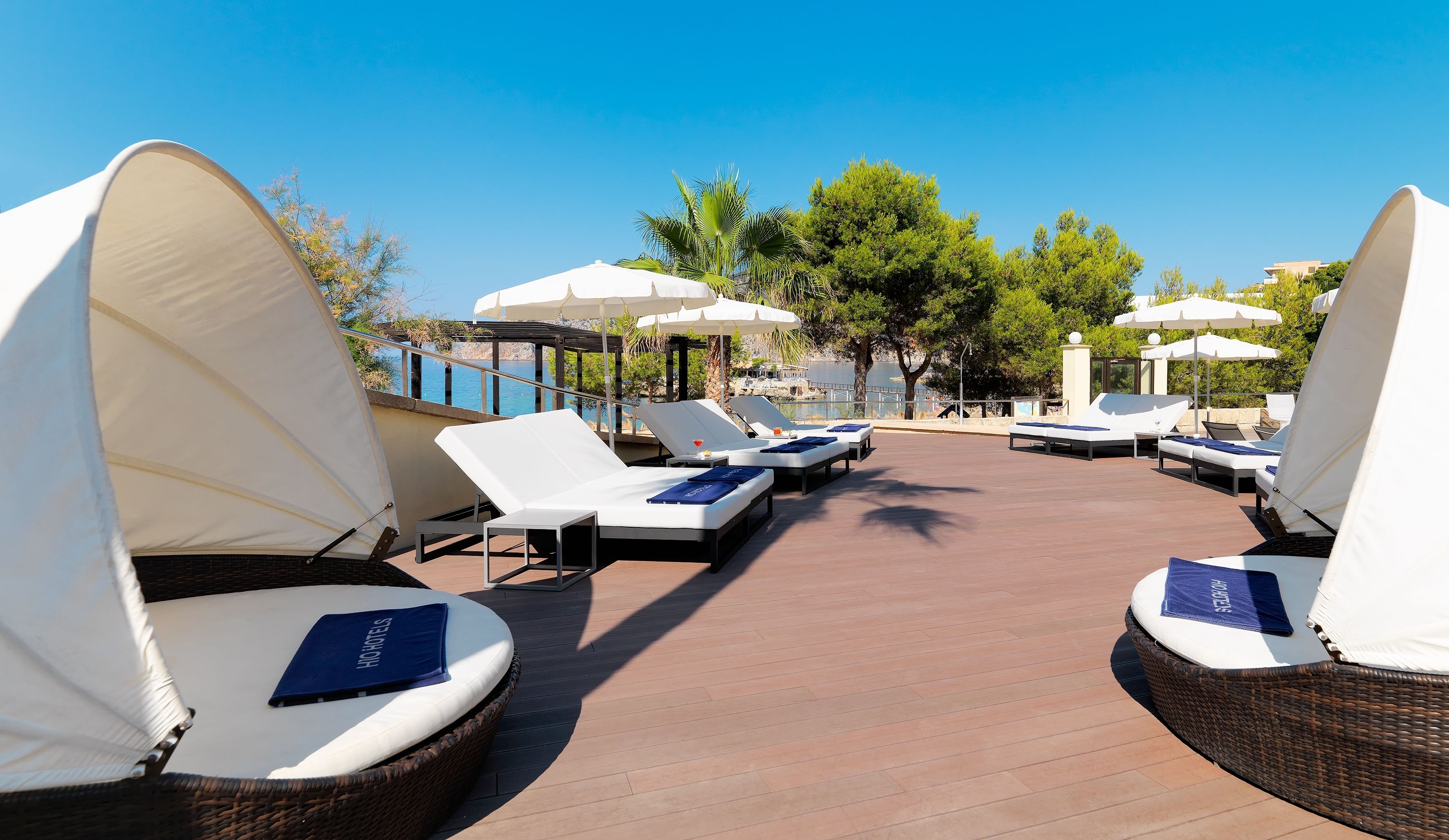 A sunlit outdoor lounge area with white lounge chairs and umbrellas, a wooden deck, and a view of the sea and greenery.