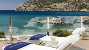Un balcón con dos sillones blancos, una barandilla de cristal y vista al mar y una colina rocosa.