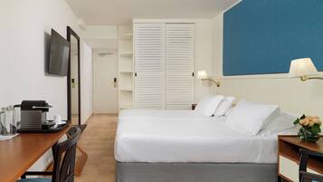 Habitación de hotel con cama, escritorio con cafetera, televisor y lámpara de pared.