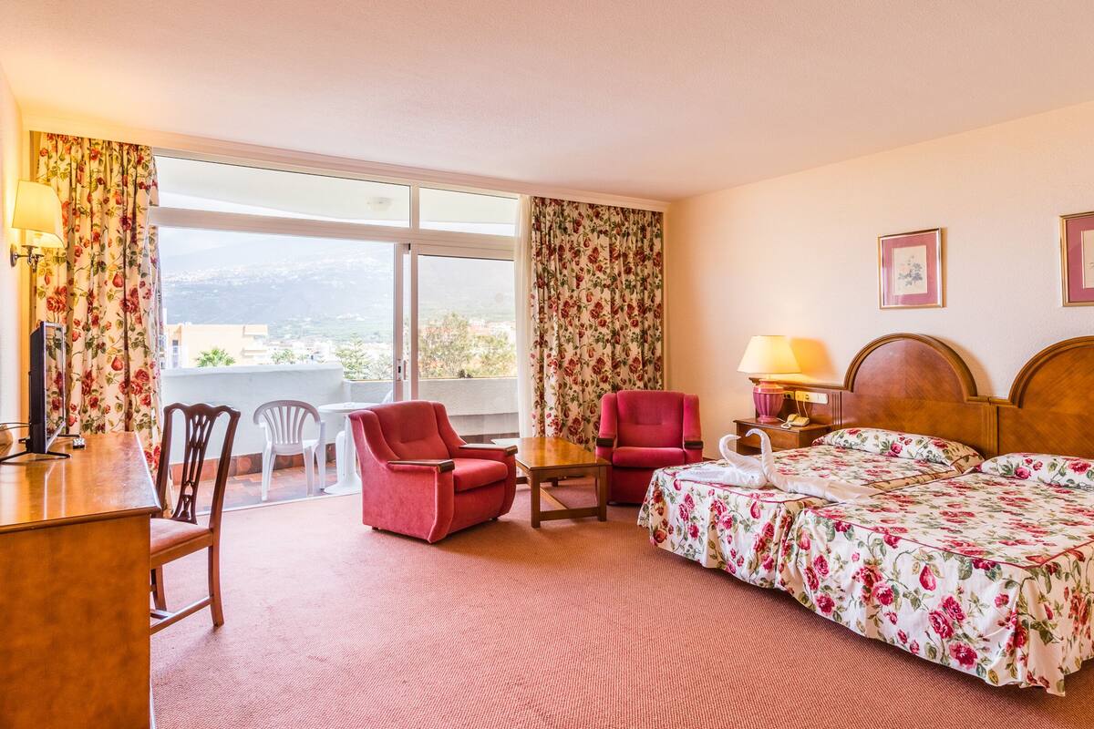Habitación de hotel con cama, dos sillas, escritorio, televisión, lámpara y ventana con cortinas.