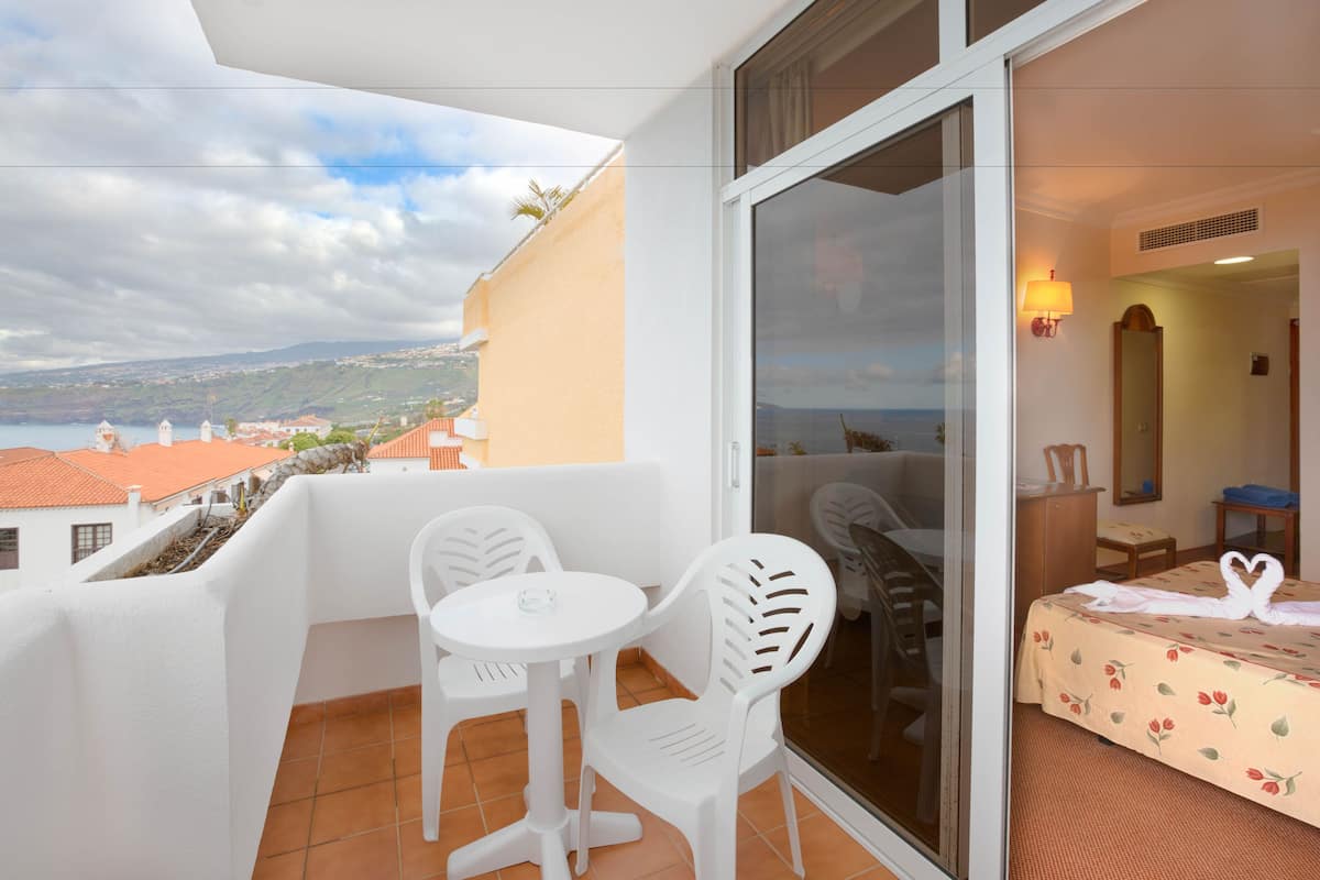 Un balcón con mesa y sillas, una cama con colcha estampada y vista al mar y edificios.