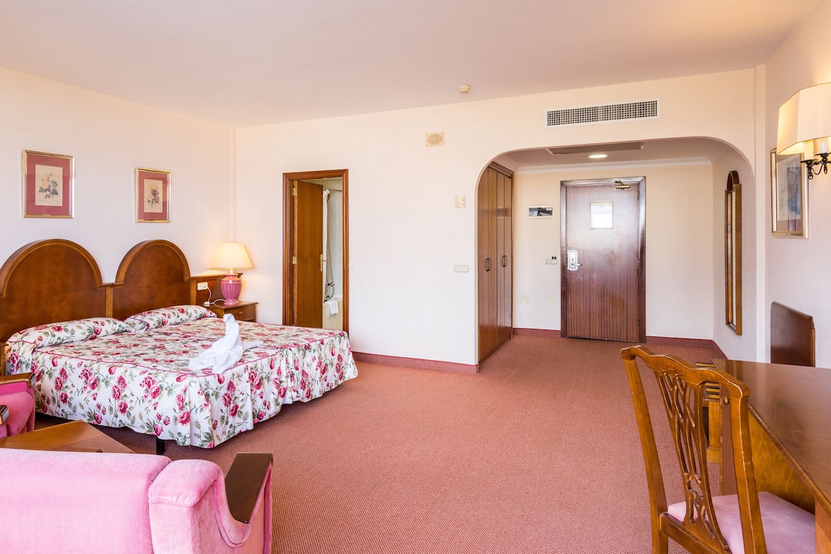 Una habitación de hotel con una cama, dos lámparas de noche, una silla, una mesa y un espejo.