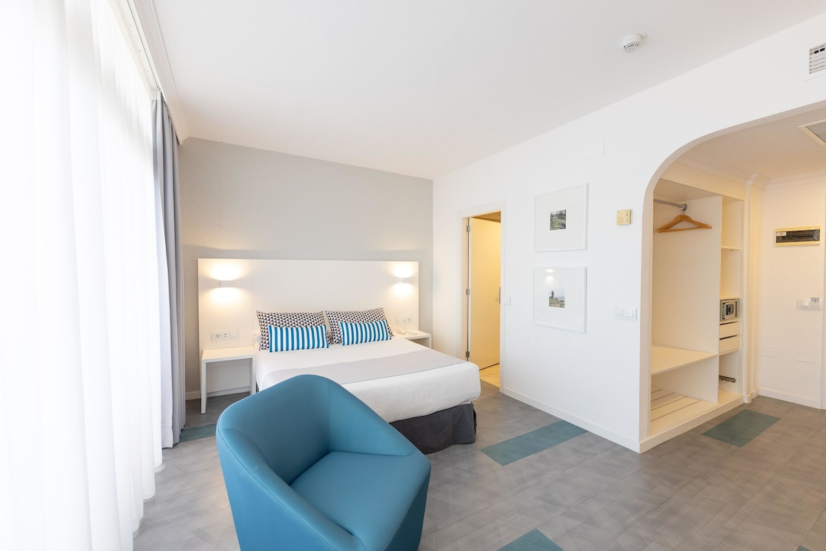 Habitación de hotel con cama, mesita de noche, lámpara y un sillón azul. Se ve una puerta que da a un baño y un armario con perchero.
