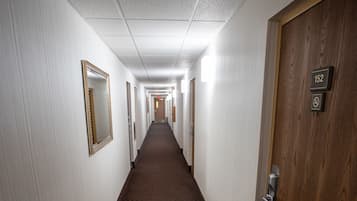 A hotel hallway with a carpeted floor, a wooden door with a number plate, and a mirror on the wall.