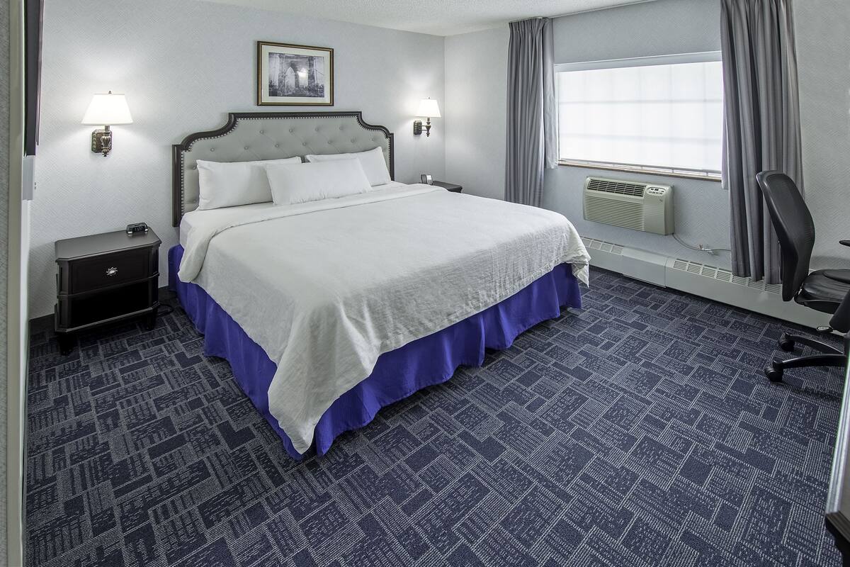 Una habitación de hotel con una cama grande, dos lámparas de noche, una mesita, una silla, un ventanal con cortinas y un aire acondicionado instalado en la pared.