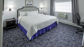 A hotel room with a large bed, two bedside lamps, a nightstand, a chair, a window with curtains, and a wall-mounted air conditioner.