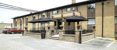A two-story building with a brick facade, outdoor seating area with umbrellas, and a red car parked in front.