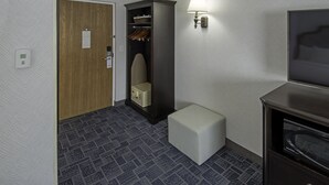 Una habitación de hotel con puerta de madera, armario con ropa colgada, lámpara de noche, un pequeño banquillo blanco y un televisor.