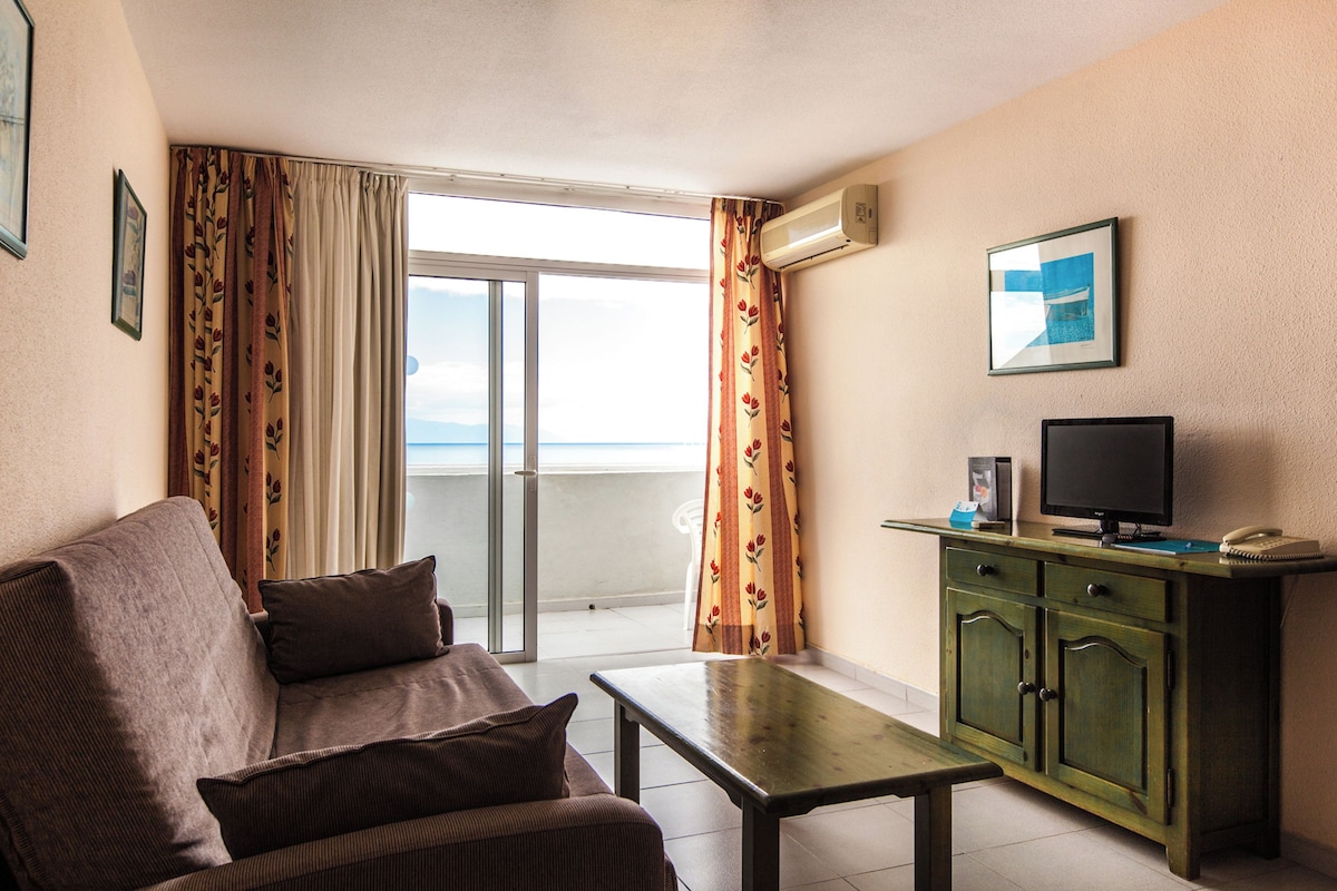 Una habitación con sofá, mesa, televisión y vista al mar a través de una ventana.