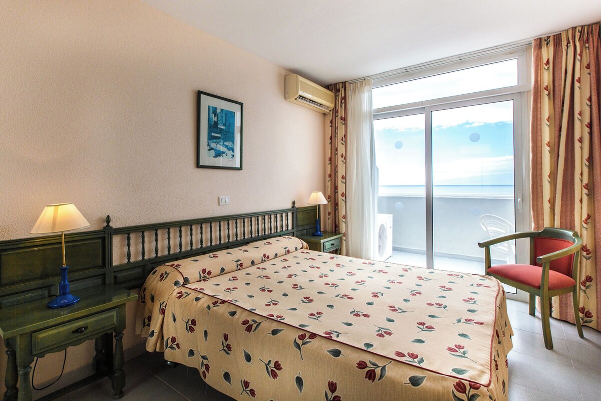 Hotellihuone, jossa on sänky, kaksi yöpöytää, tuoli, ikkuna verhoilla, ilmastointilaite ja seinällä oleva kehystetty kuva.