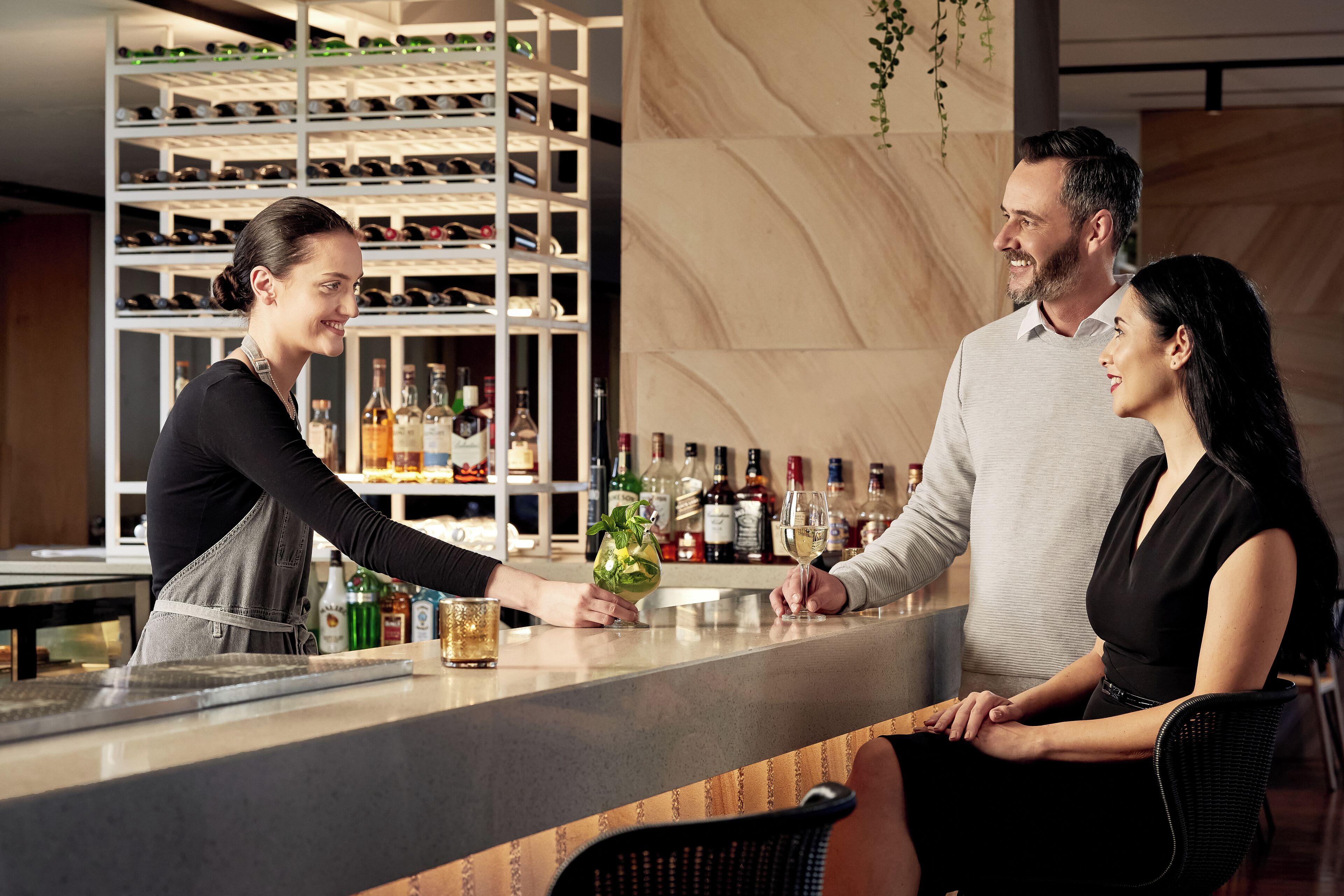 A bar area with a bartender serving drinks to guests. There are shelves with bottles in the background.