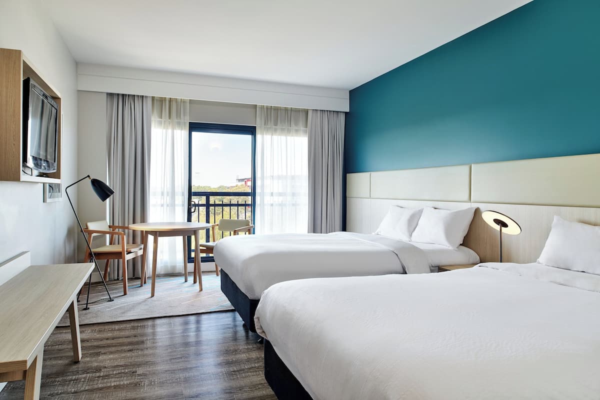 A hotel room with two beds, a desk, a chair, and a view of the outdoors.