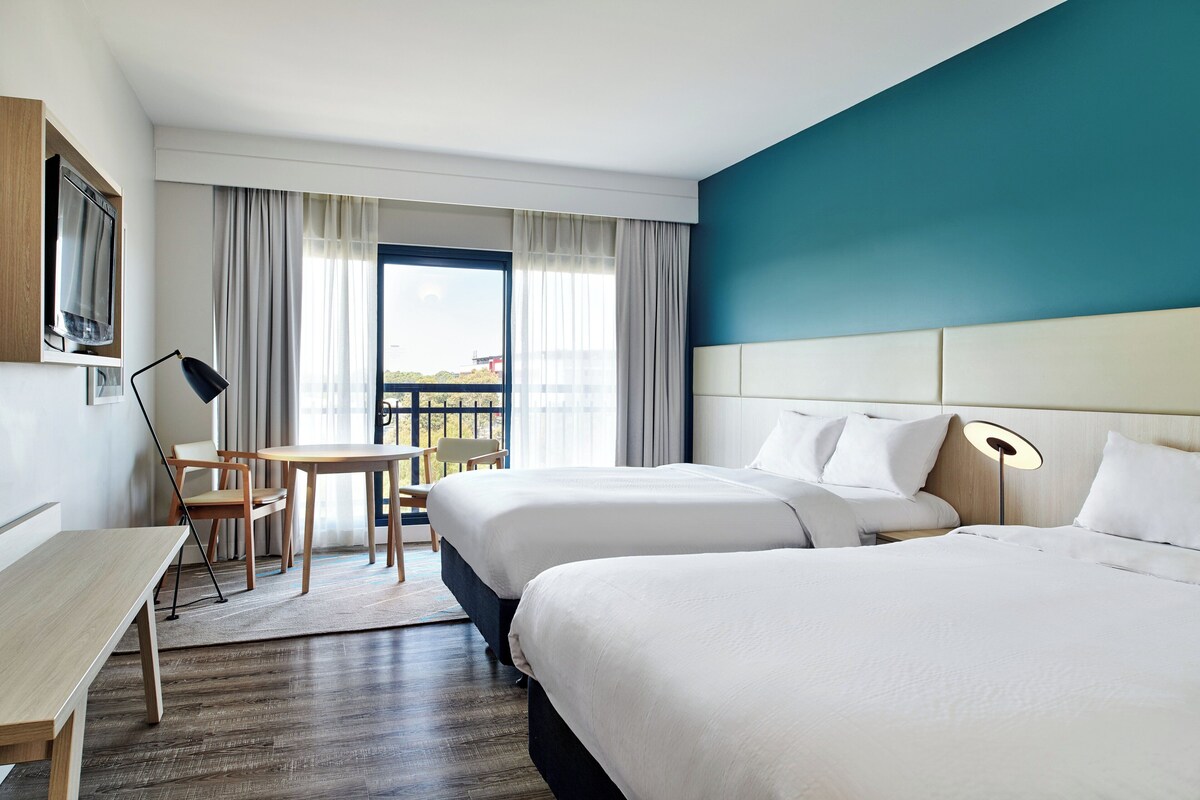 A hotel room with two beds, a desk, a chair, and a view of the outdoors.