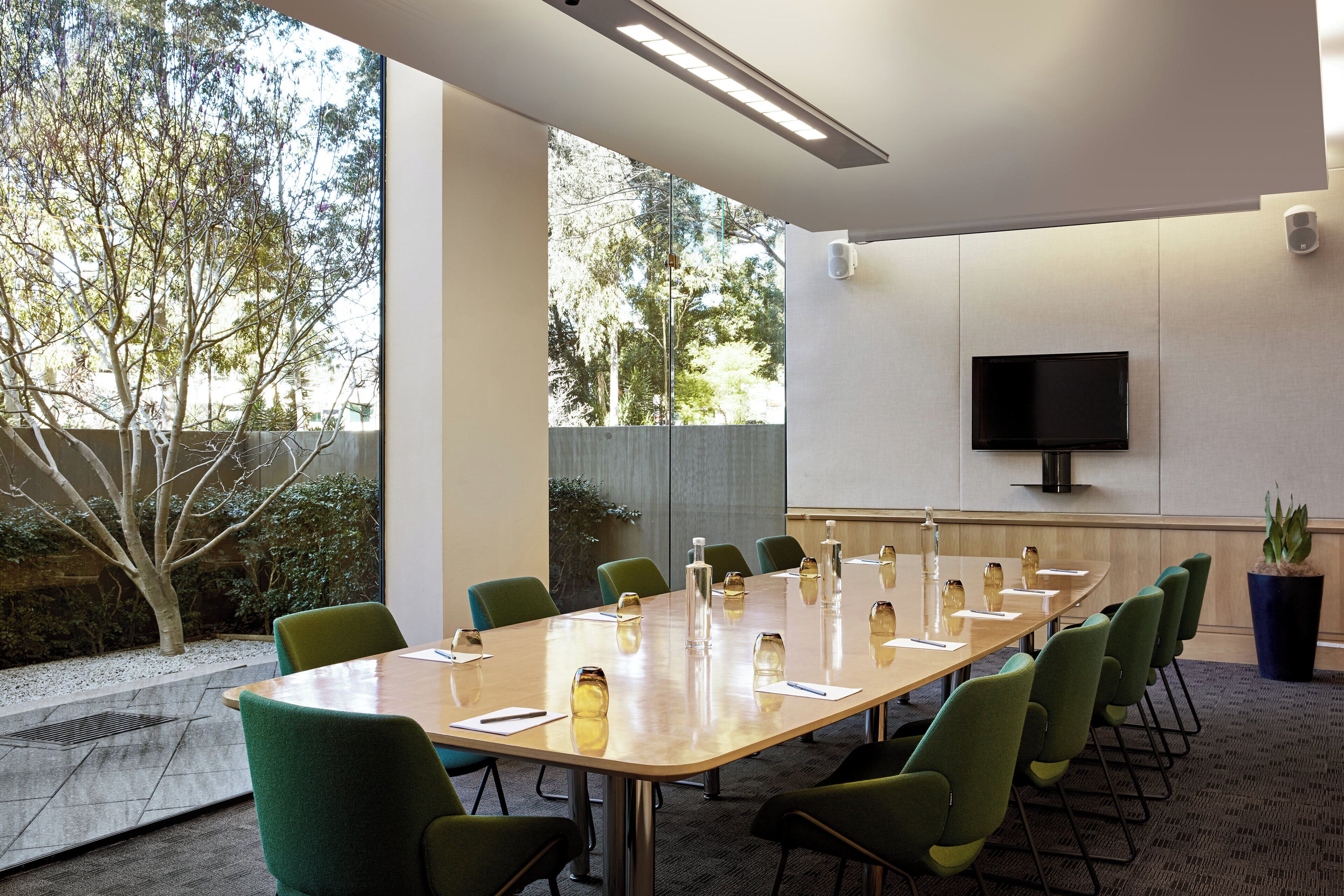 Un salón de conferencias con una mesa larga, sillas verdes, un televisor de pantalla plana en la pared y una planta en maceta.