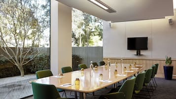 Un salón de conferencias con una mesa larga, sillas verdes, un televisor de pantalla plana en la pared y una planta en maceta.