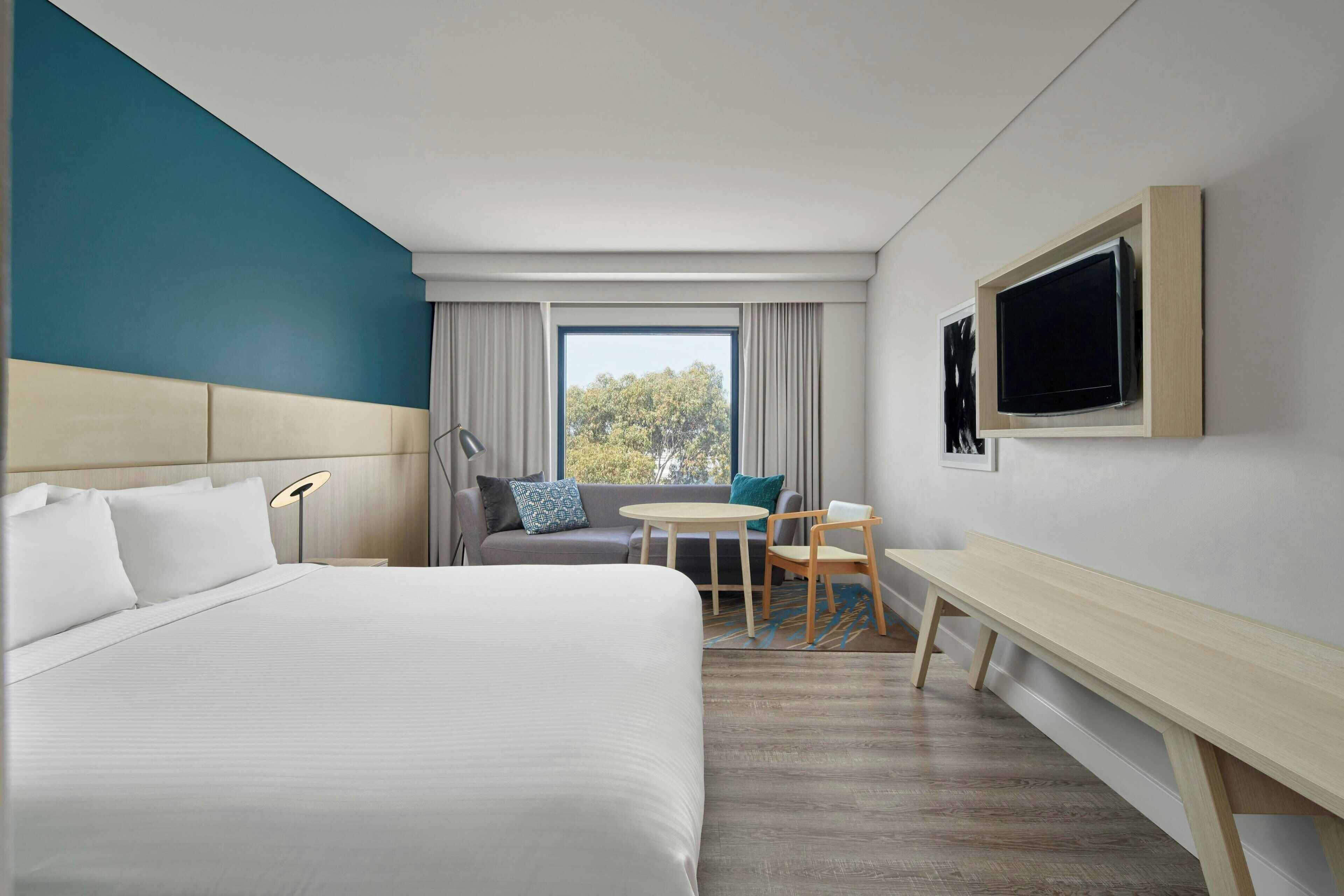 A hotel room with a large bed, a desk, a chair, a television, and a window with a view of trees.