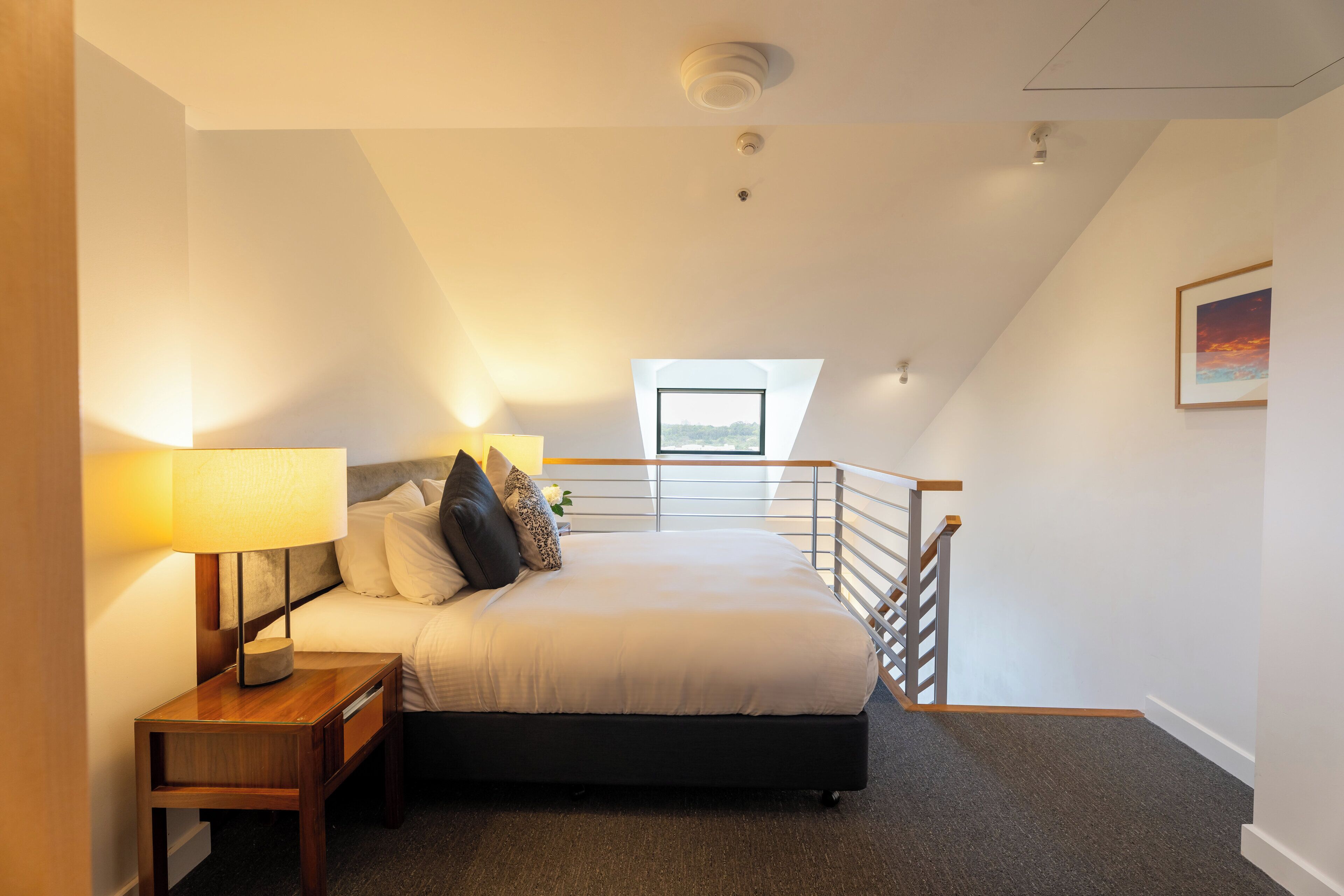A bedroom with a bed, nightstand, lamp, and a view of the city through a skylight.