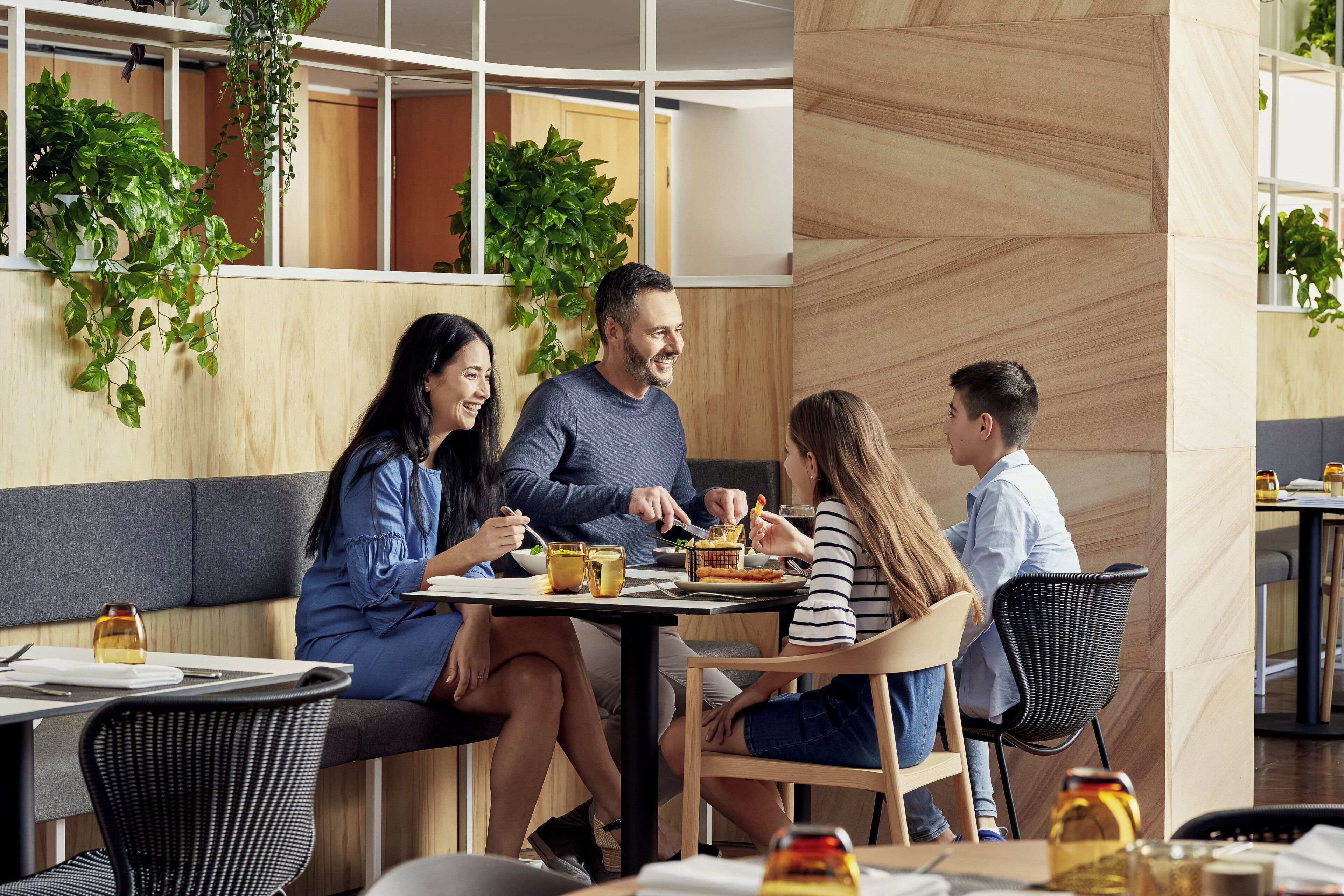 A modern dining area with wooden walls, green plants, and a mix of seating arrangements including a bench and chairs.