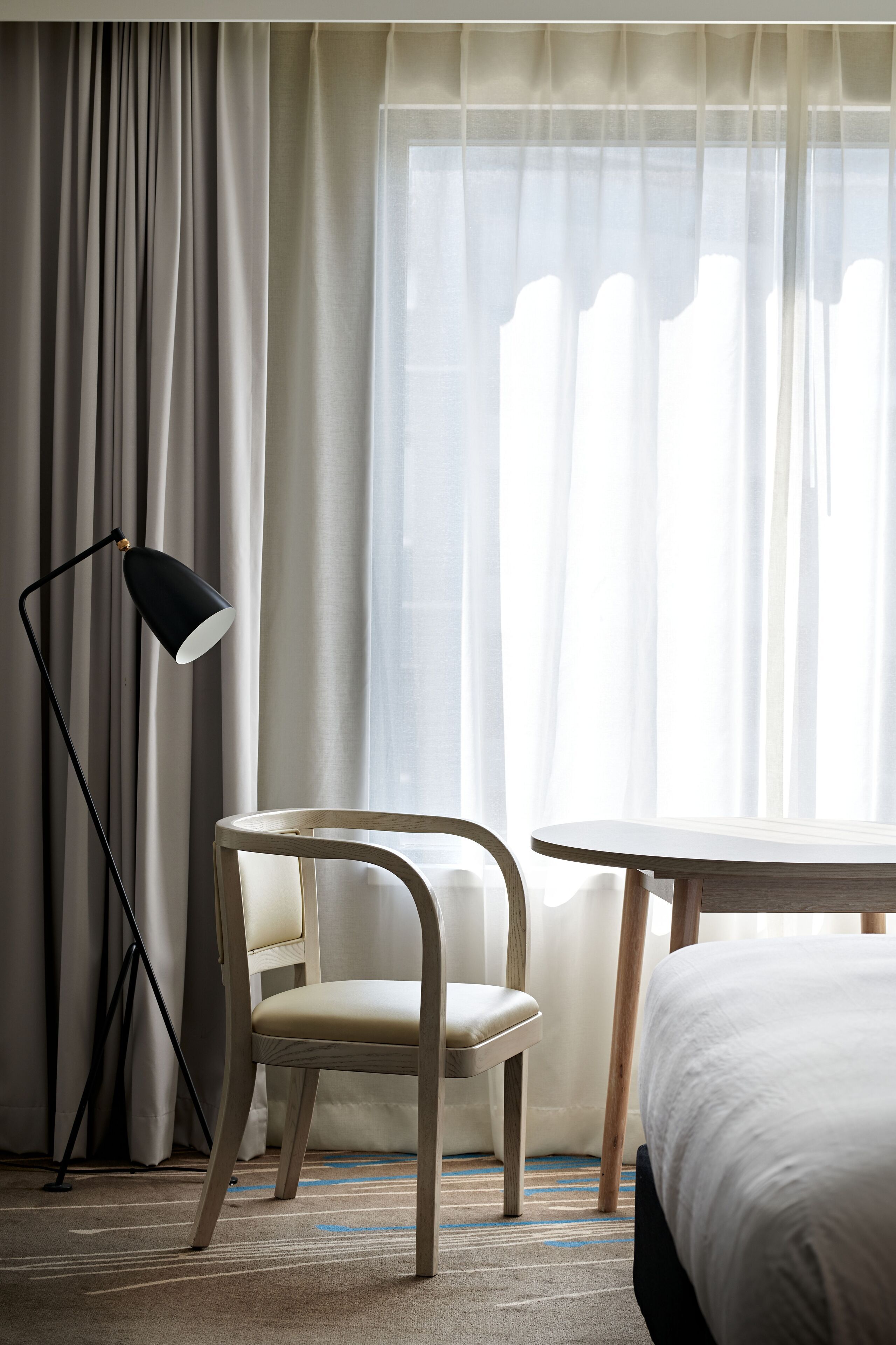 Ett hotellrum med en säng, en stol, ett litet bord, en golvlampa och tunna, genomskinliga gardiner.