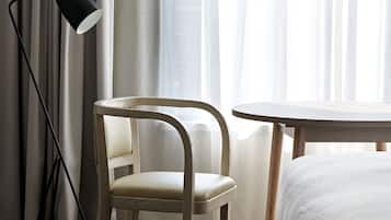 Quarto de hotel com cama, cadeira, mesa pequena, abajur e cortinas translúcidas.