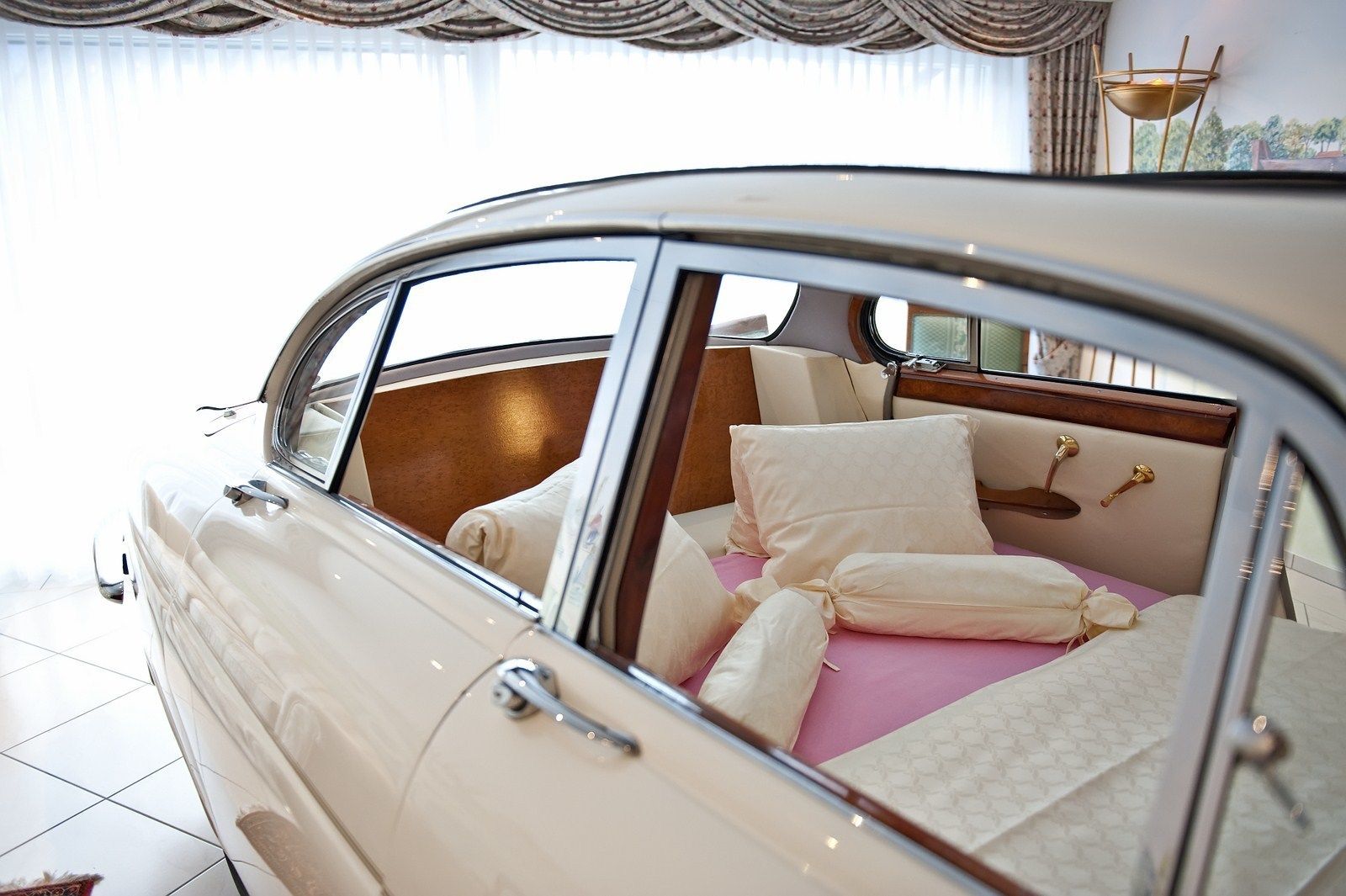 Un coche clásico con techo descubierto, que muestra un interior lujoso con cama, almohadas y una mesita de noche.