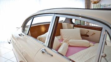 A vintage car with a convertible top, revealing a luxurious interior with a bed, pillows, and a nightstand.