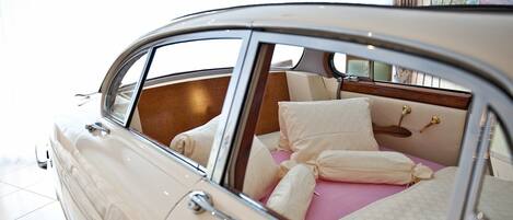 Un coche clásico con techo descubierto, que muestra un interior lujoso con cama, almohadas y una mesita de noche.