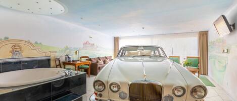 Un coche antiguo en una habitación amplia con una bañera grande, una zona de comedor y un mural en la pared.