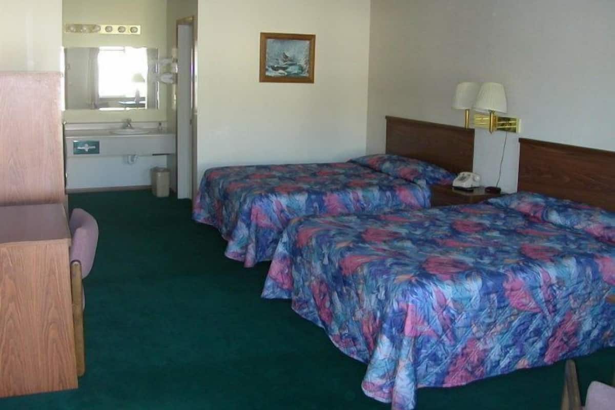 A hotel room with two beds, a desk, a chair, a mirror, and a small table with a lamp.