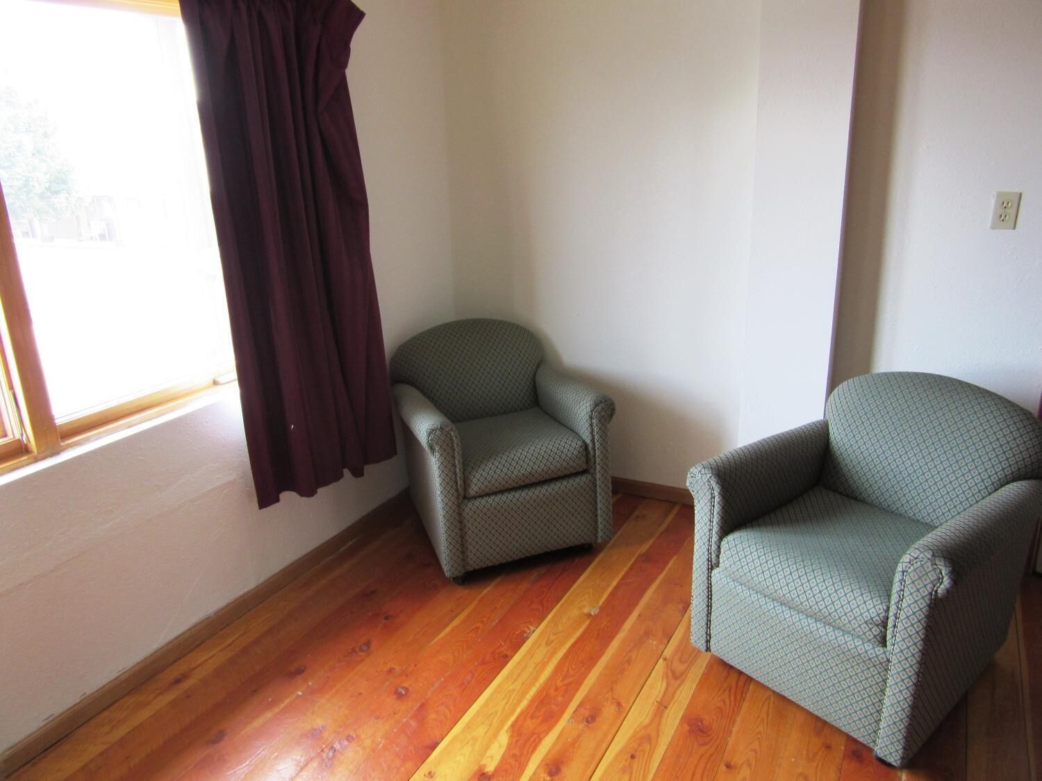 Una habitación con suelo de madera, dos sillones, un ventanal con cortina y una pared blanca lisa.