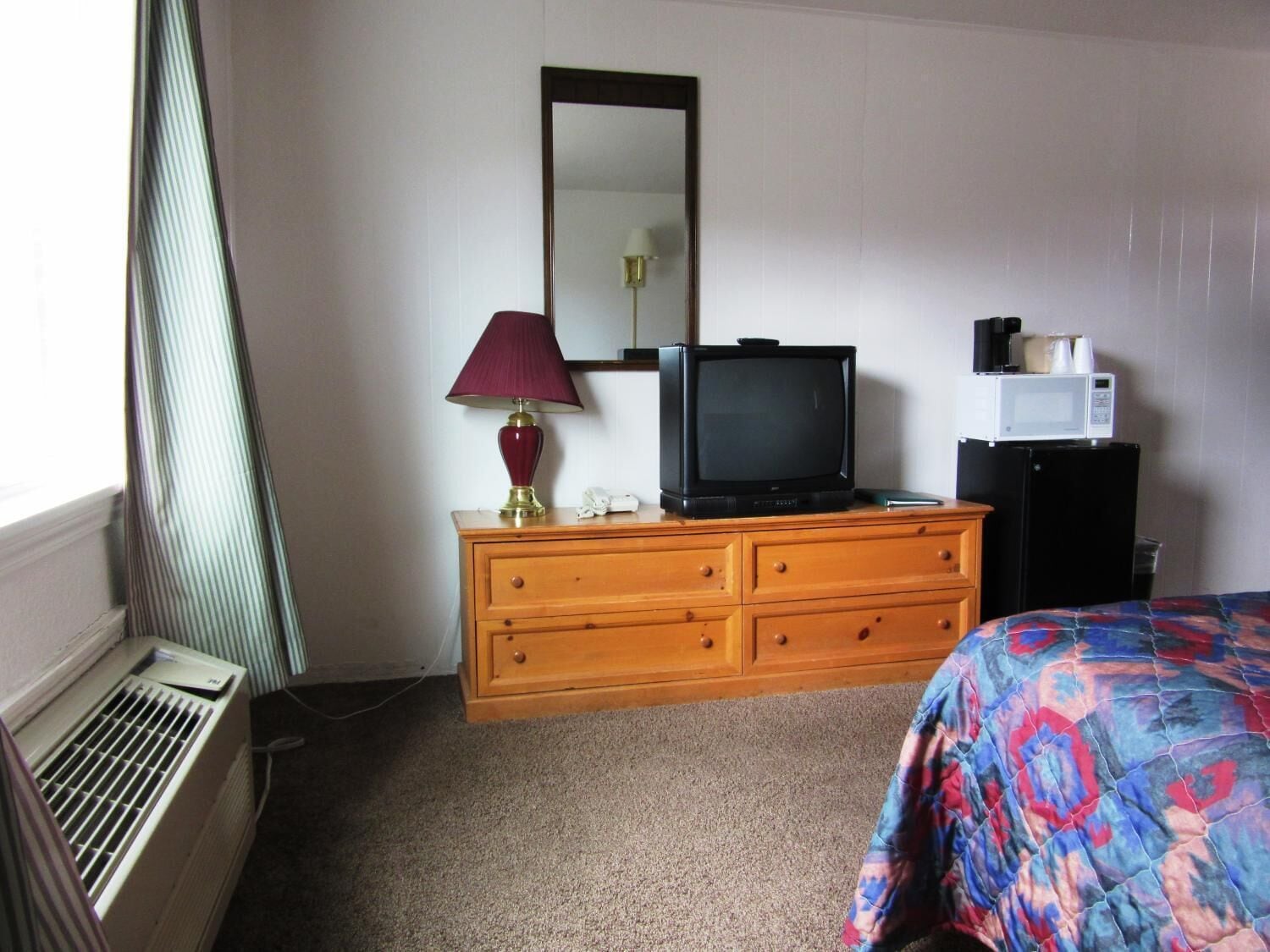A hotel room with a wooden dresser, a television, a microwave, and a bed with a colorful blanket.