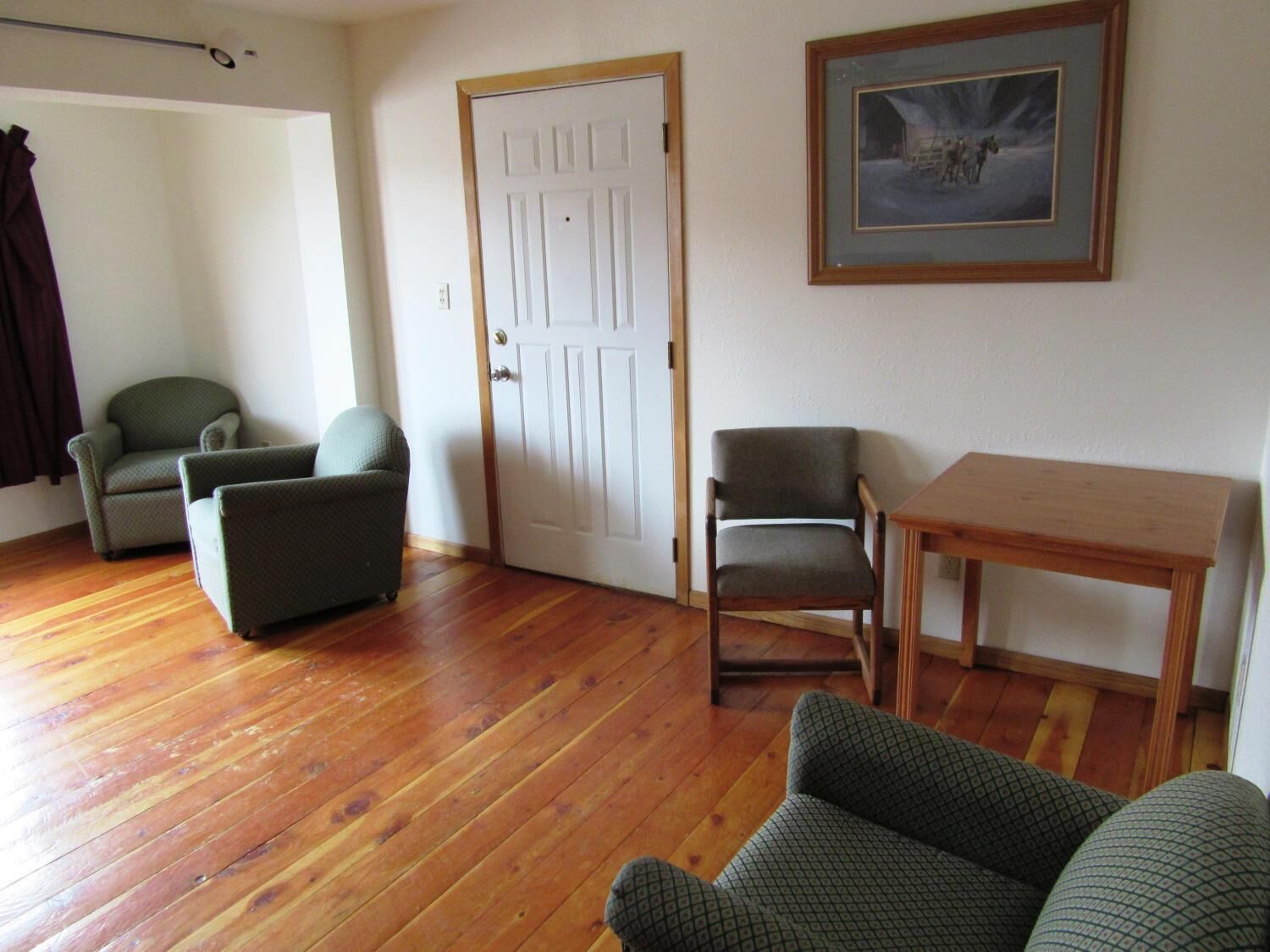 Une pièce avec un plancher en bois, une porte blanche, deux fauteuils, une petite table et un tableau encadré au mur.