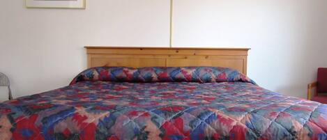 A bed with a patterned quilt, a wooden headboard, a wall-mounted lamp, and a framed picture on the wall.