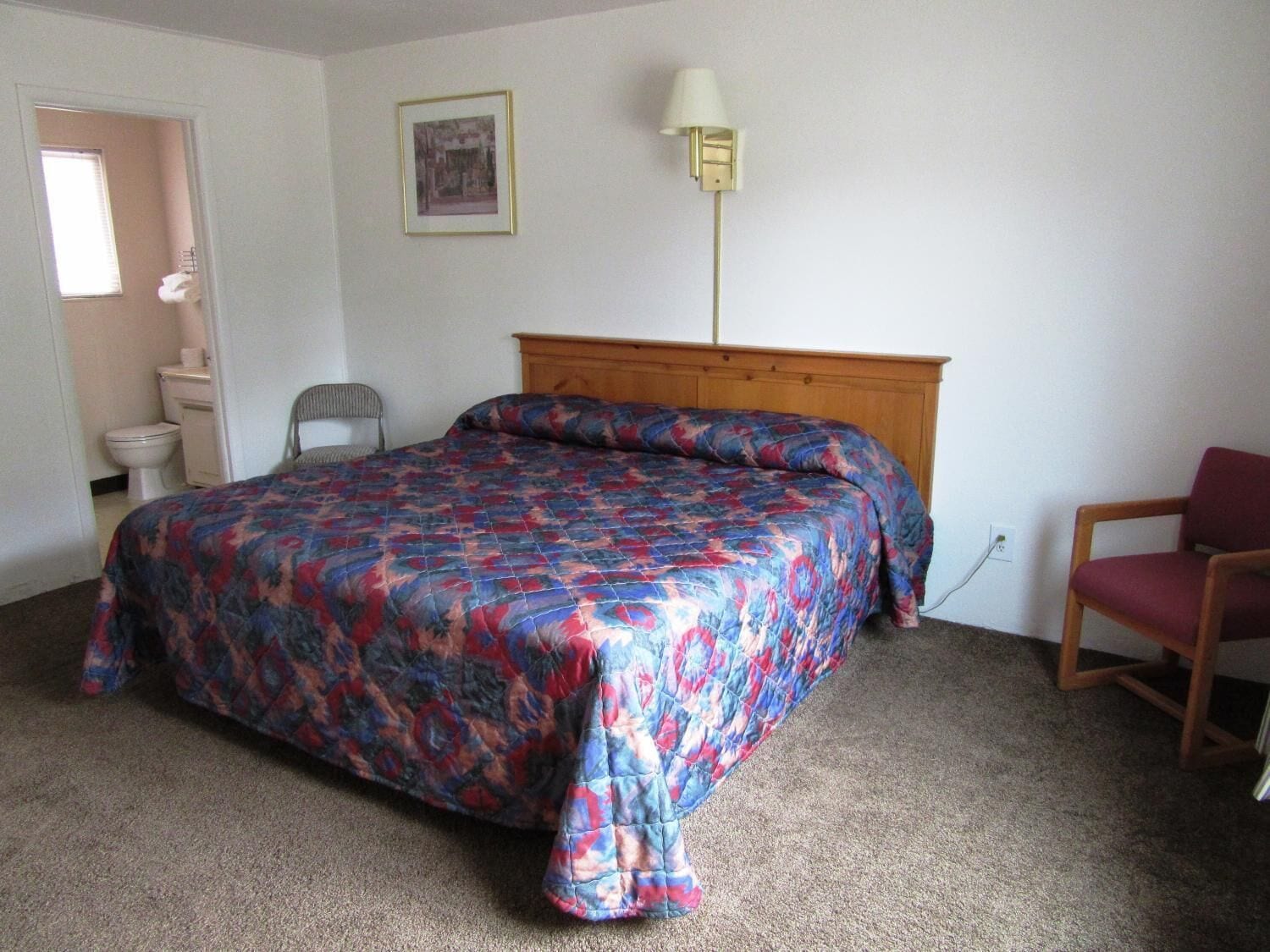A bedroom with a bed, a chair, a bedside table, a lamp, and a framed picture on the wall.